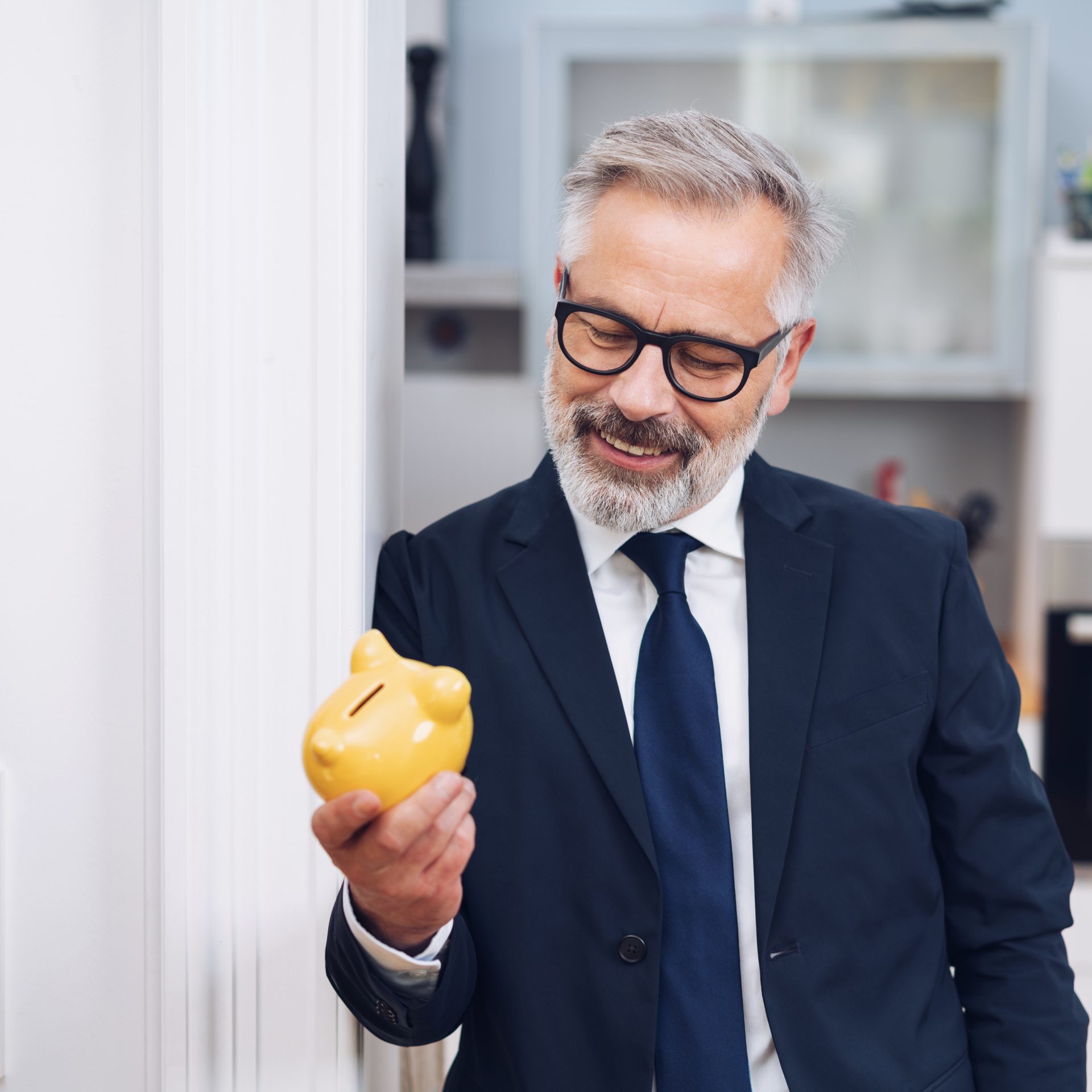 Successful businessman looking at a yellow handheld piggy bank with a pleased smile as he leans against a door frame at home